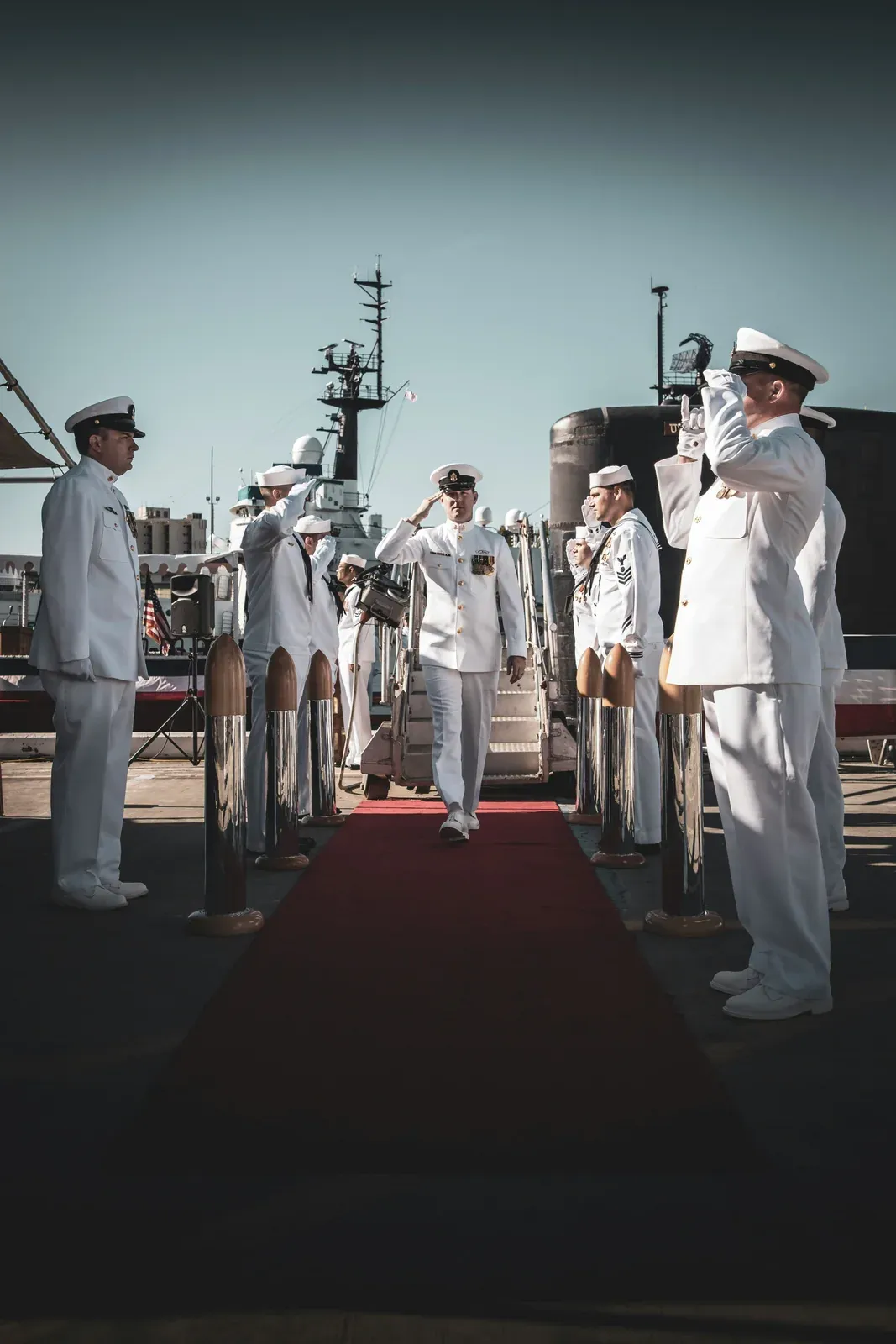 US Navy petty officers in dress white uniforms salute as an officer crosses the pier